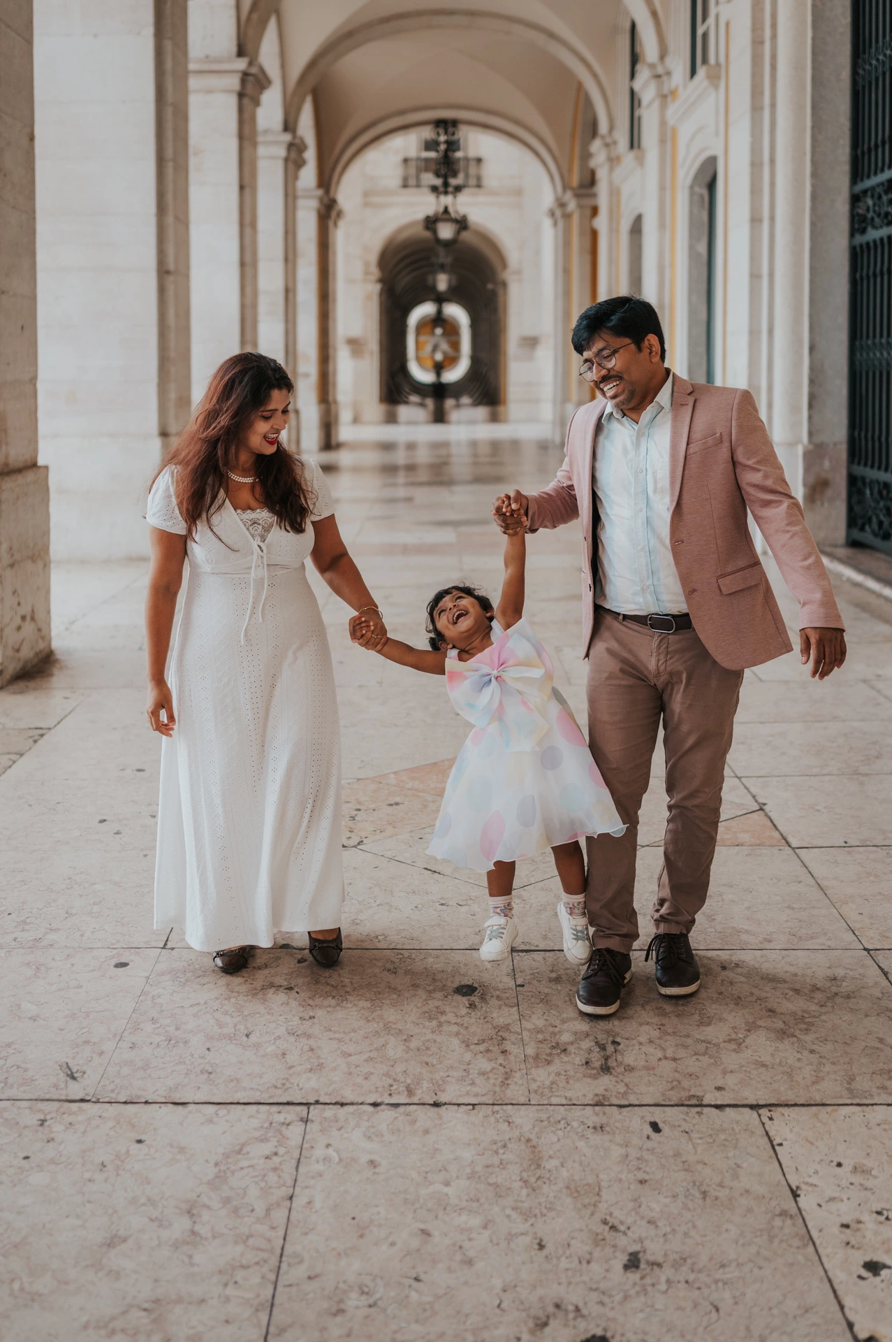 The parents holding their daughter’s hands as they walk together beneath the arcade during their family birthday photoshoot in Lisbon.