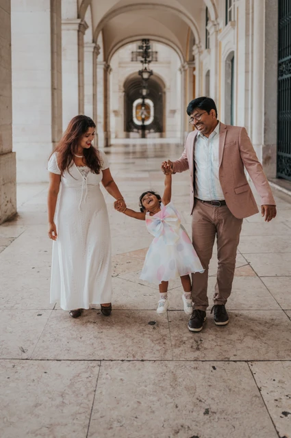 A playful family walk beneath the arches | Lisbon family birthday photoshoot The parents holding their daughter’s hands as they walk together beneath the arcade during their family birthday photoshoot in Lisbon.