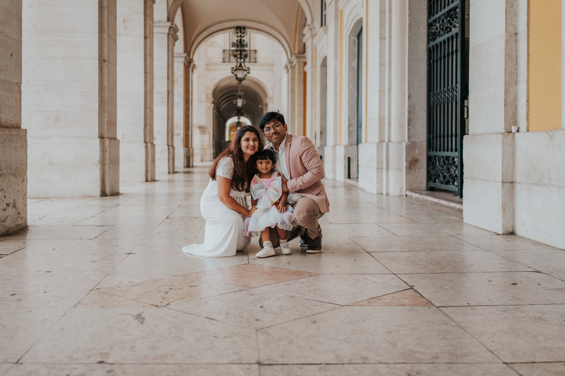 A family portrait at their daughter’s height | Lisbon family birthday photoshoot The parents crouching beside their daughter for a family portrait beneath the arcade during their family birthday photoshoot in Lisbon.