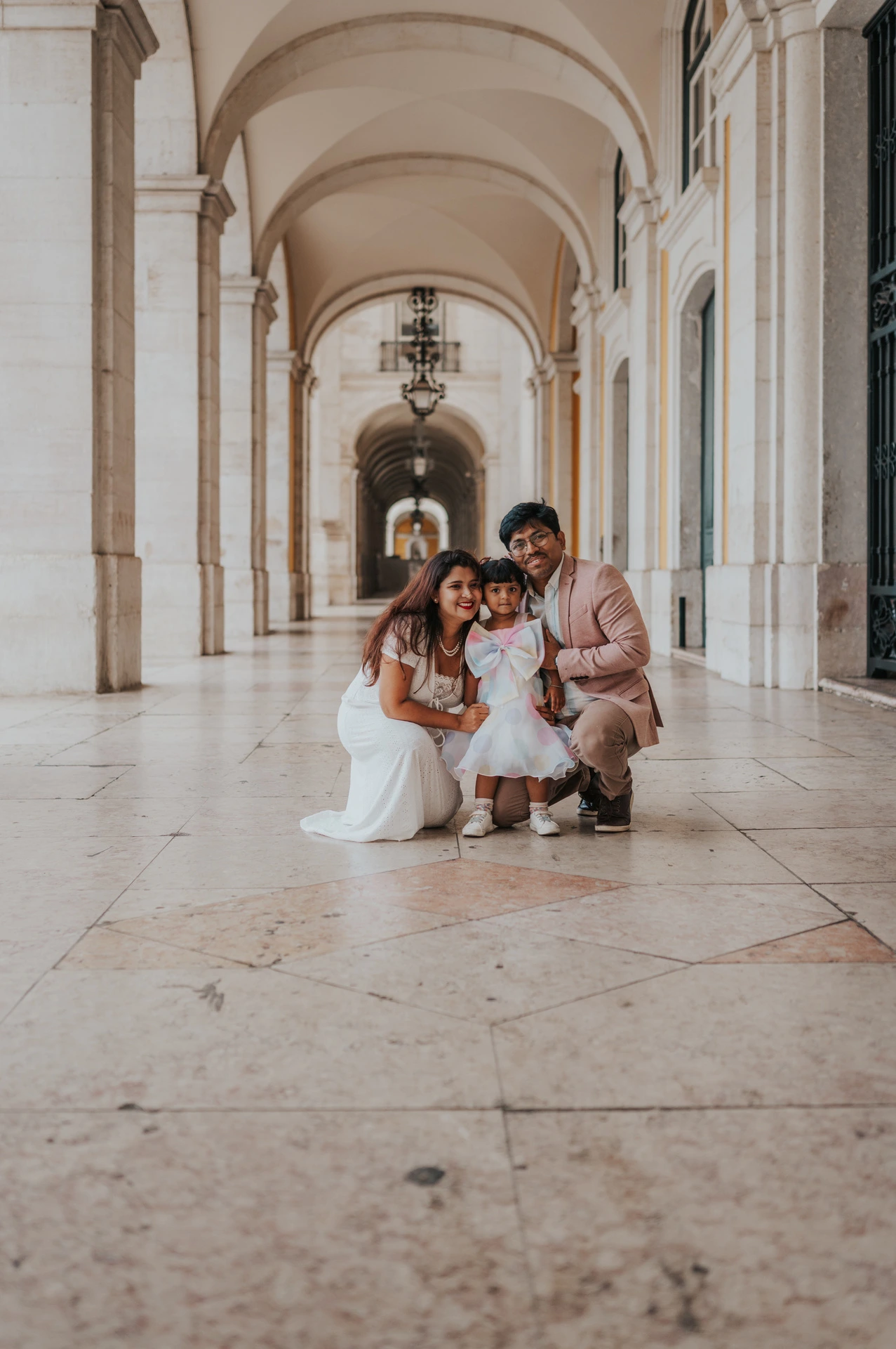 The parents crouching beside their daughter for a family portrait beneath the arcade during their family birthday photoshoot in Lisbon.
