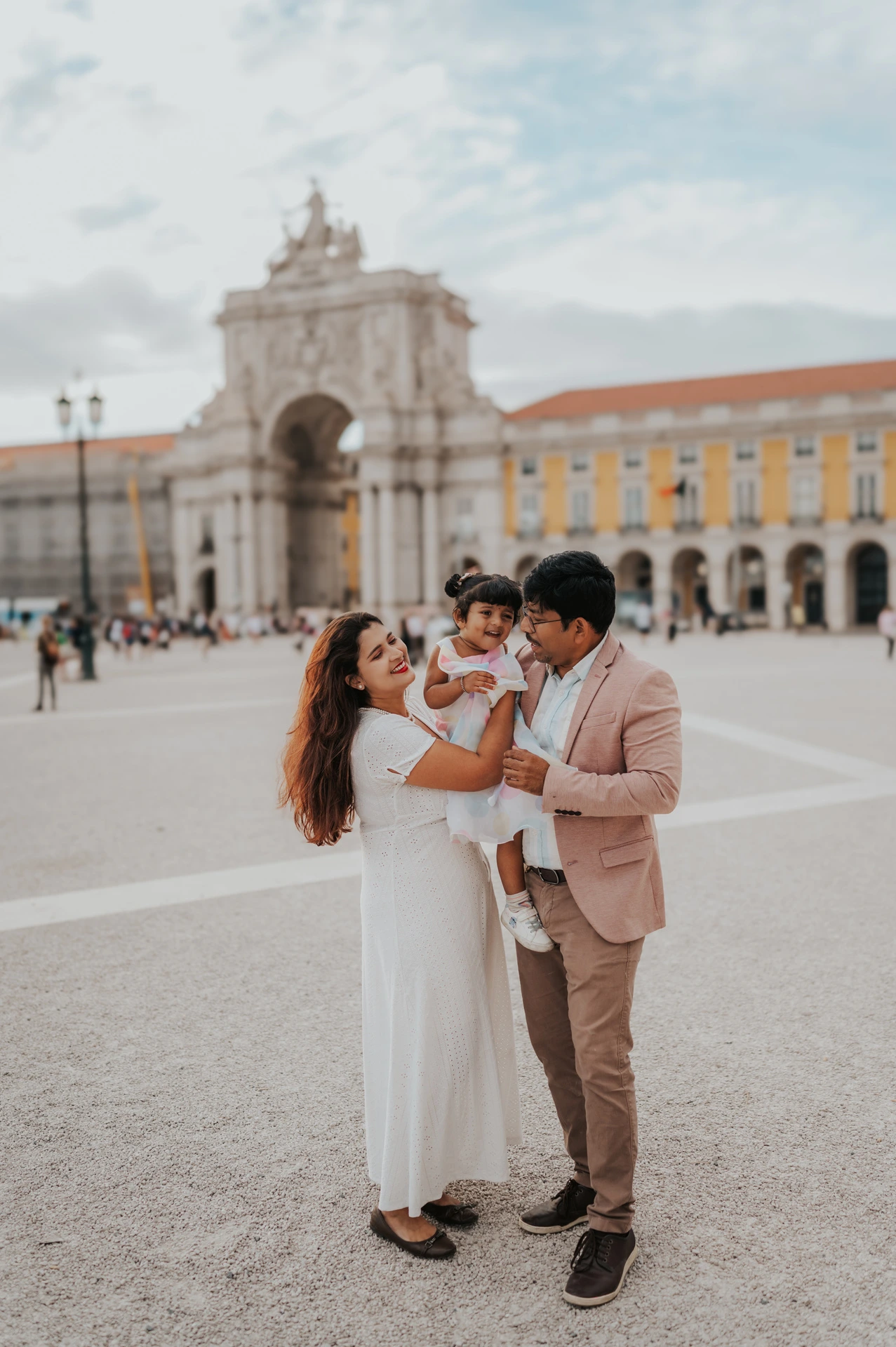 The parents holding their daughter close together in front of the square’s monumental arch during their family birthday photoshoot in Lisbon.