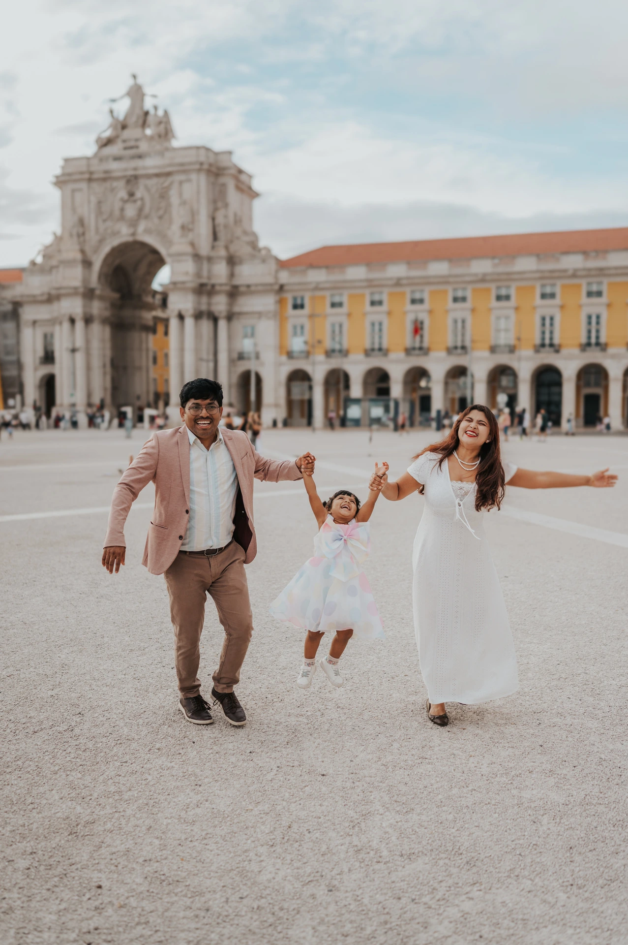 The parents holding their daughter’s hands as they walk together in front of Praça do Comércio’s arch during their family birthday photoshoot in Lisbon.