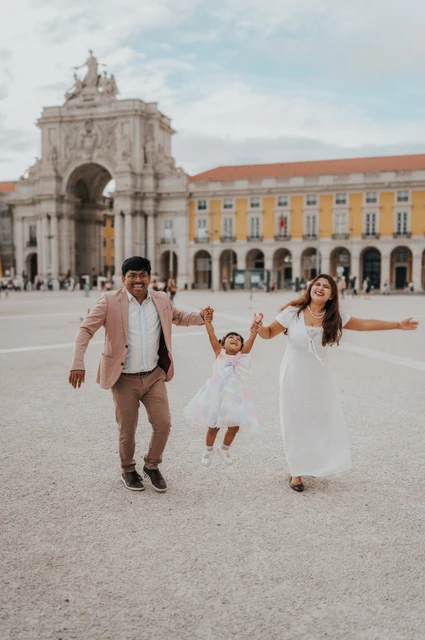 Hand in hand across the open square | Lisbon family birthday photoshoot The parents holding their daughter’s hands as they walk together in front of Praça do Comércio’s arch during their family birthday photoshoot in Lisbon.