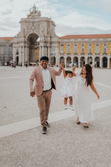 Hand in hand across the open square | Lisbon family birthday photoshoot The parents holding their daughter’s hands as they walk together in front of Praça do Comércio’s arch during their family birthday photoshoot in Lisbon.