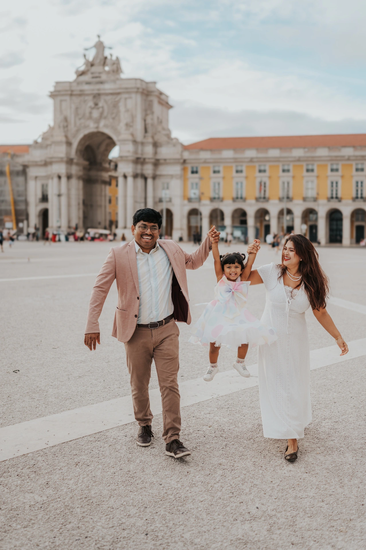 The parents holding their daughter’s hands as they walk together in front of Praça do Comércio’s arch during their family birthday photoshoot in Lisbon.