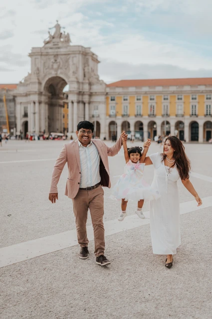 The parents holding their daughter’s hands as they walk together in front of Praça do Comércio’s arch during their family birthday photoshoot in Lisbon.