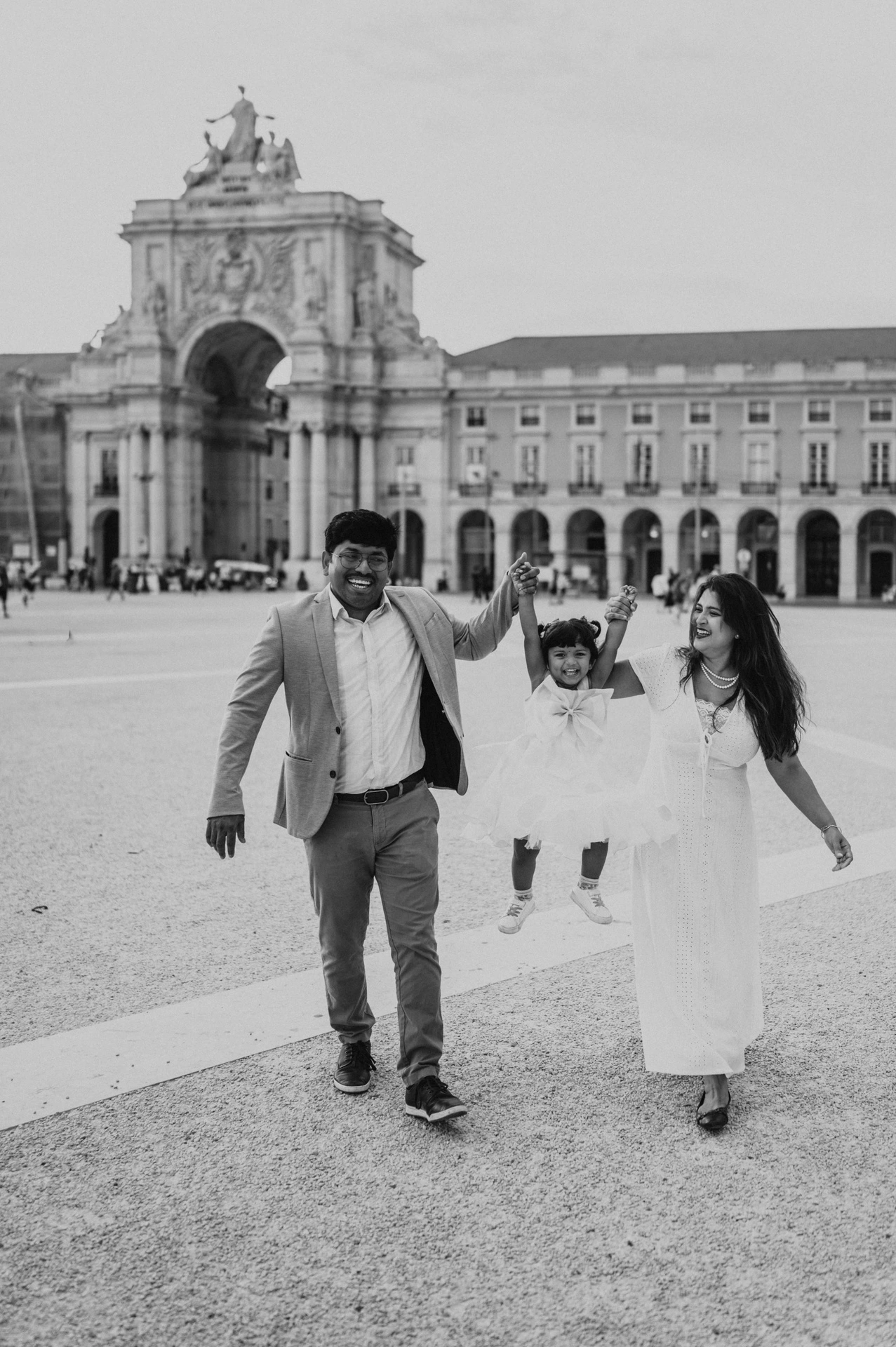 Black-and-white photograph: the parents holding their daughter’s hands as they walk together in front of Praça do Comércio’s arch during their family birthday photoshoot in Lisbon.