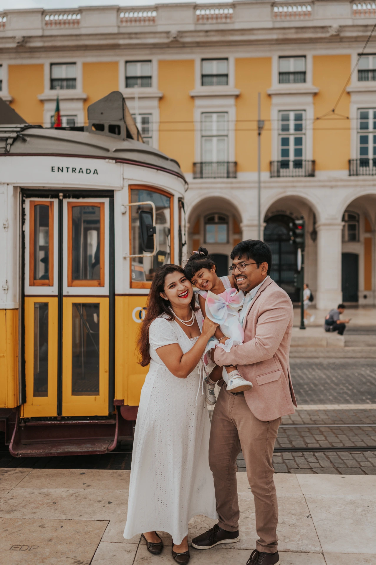The parents holding their smiling daughter together beside a yellow Lisbon tram during their family birthday photoshoot in Lisbon.