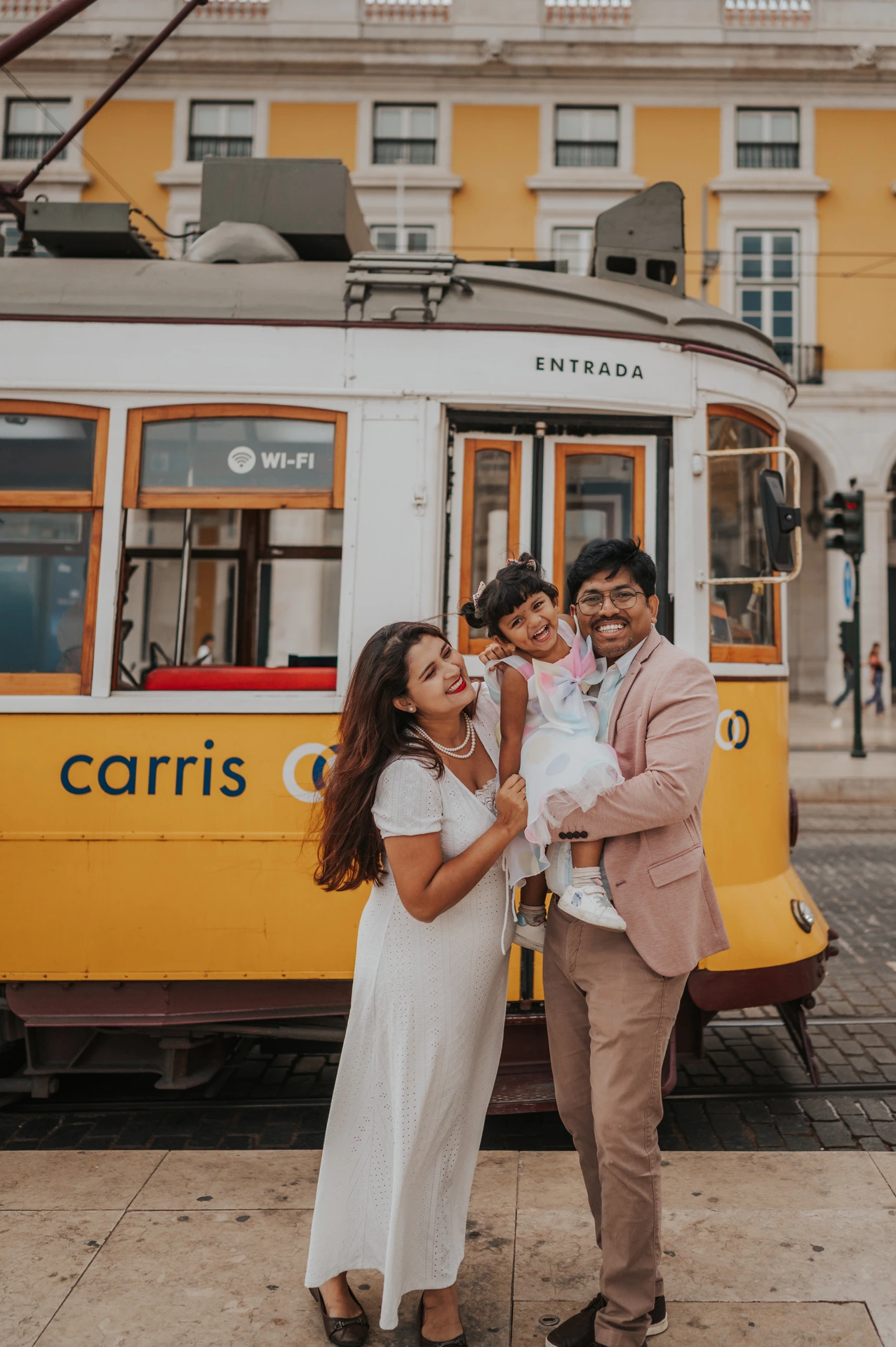 The parents holding their smiling daughter together beside a yellow Lisbon tram during their family birthday photoshoot in Lisbon.