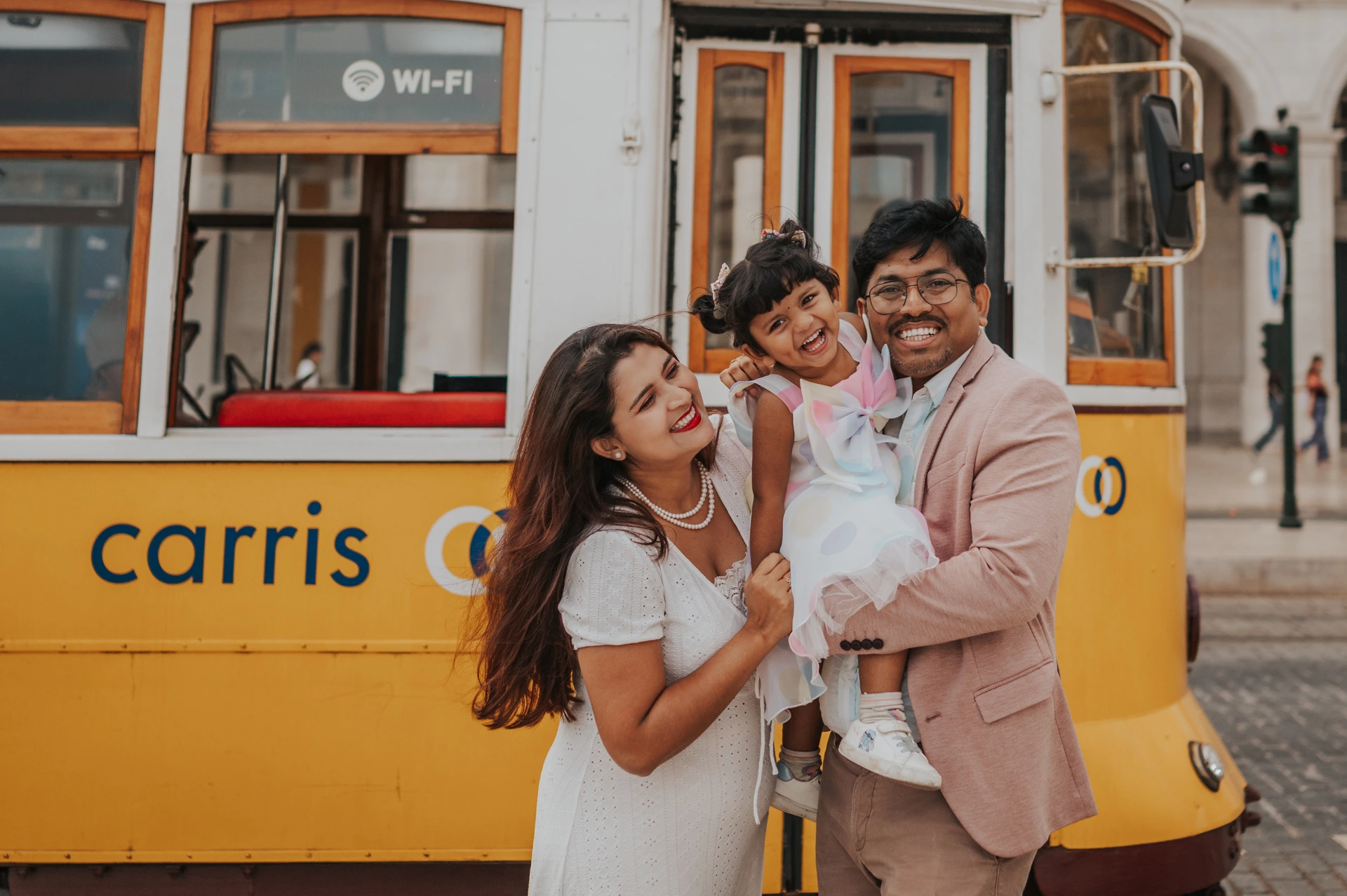 A family birthday portrait beside the yellow tram | Lisbon family birthday photoshoot The parents holding their smiling daughter together beside a yellow Lisbon tram during their family birthday photoshoot in Lisbon.