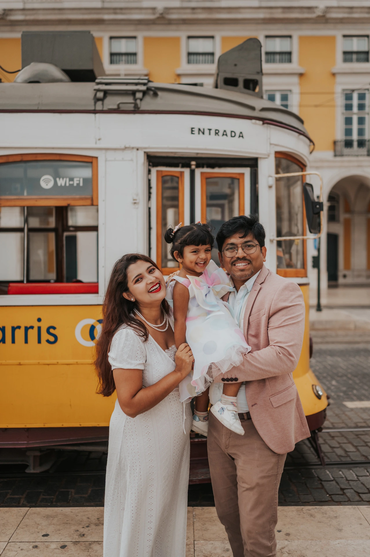 The parents holding their smiling daughter together beside a yellow Lisbon tram during their family birthday photoshoot in Lisbon.