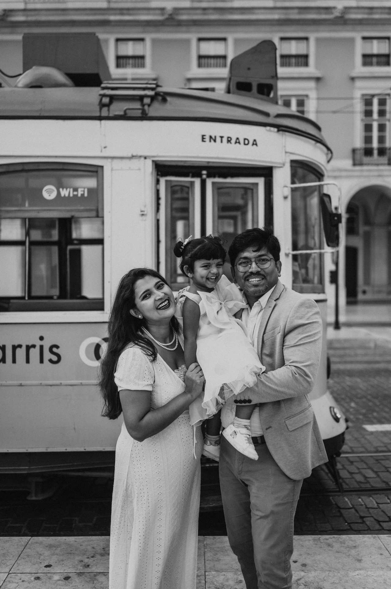 Black-and-white photograph: the parents holding their smiling daughter together beside a yellow Lisbon tram during their family birthday photoshoot in Lisbon.