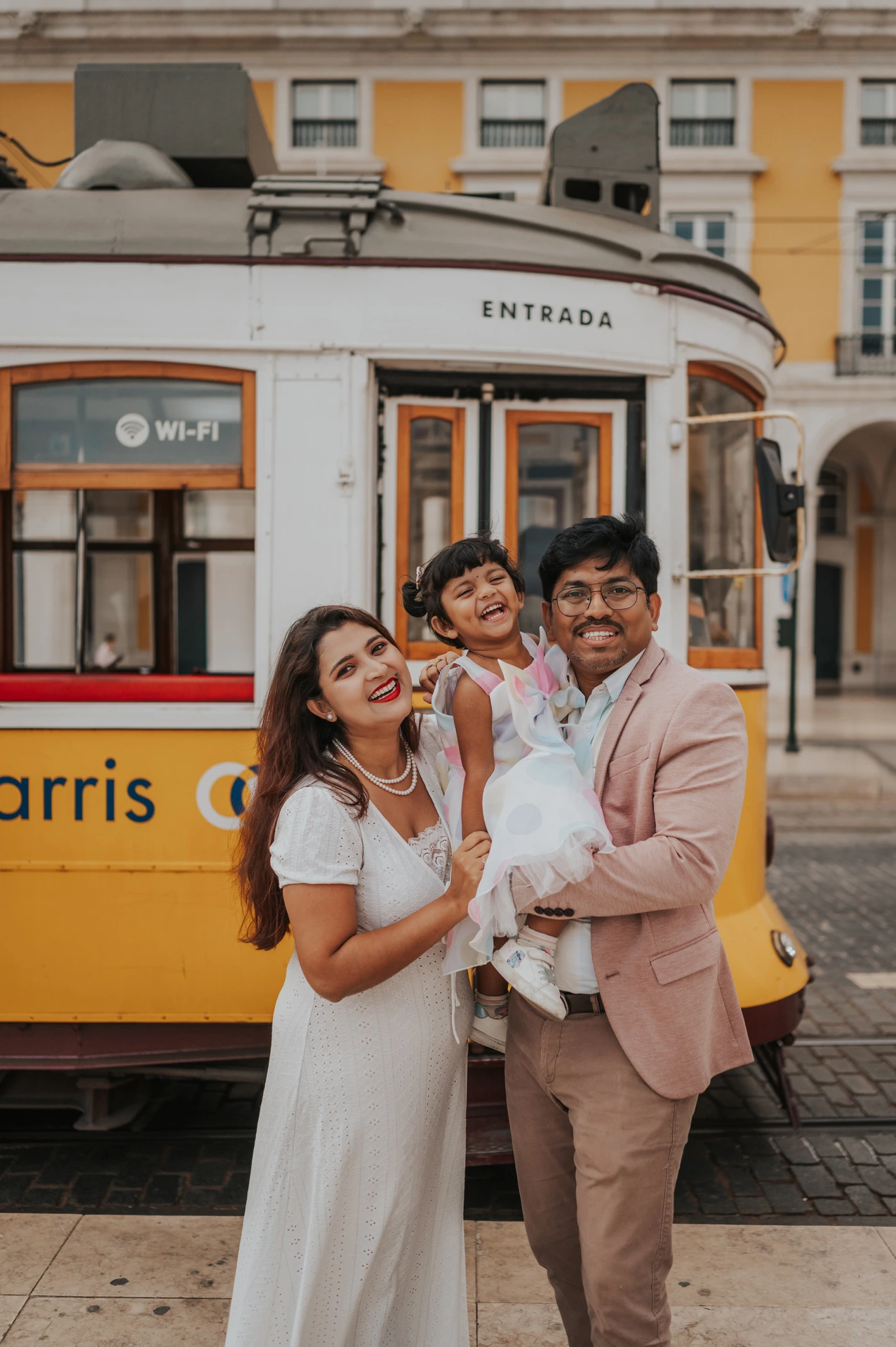 The parents holding their smiling daughter together beside a yellow Lisbon tram during their family birthday photoshoot in Lisbon.