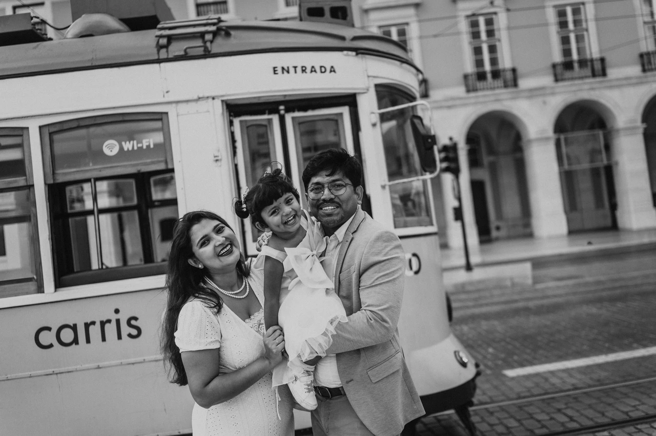A family birthday portrait beside the yellow tram in black and white | Lisbon family birthday photoshoot Black-and-white photograph: the parents holding their smiling daughter together beside a yellow Lisbon tram during their family birthday photoshoot in Lisbon.