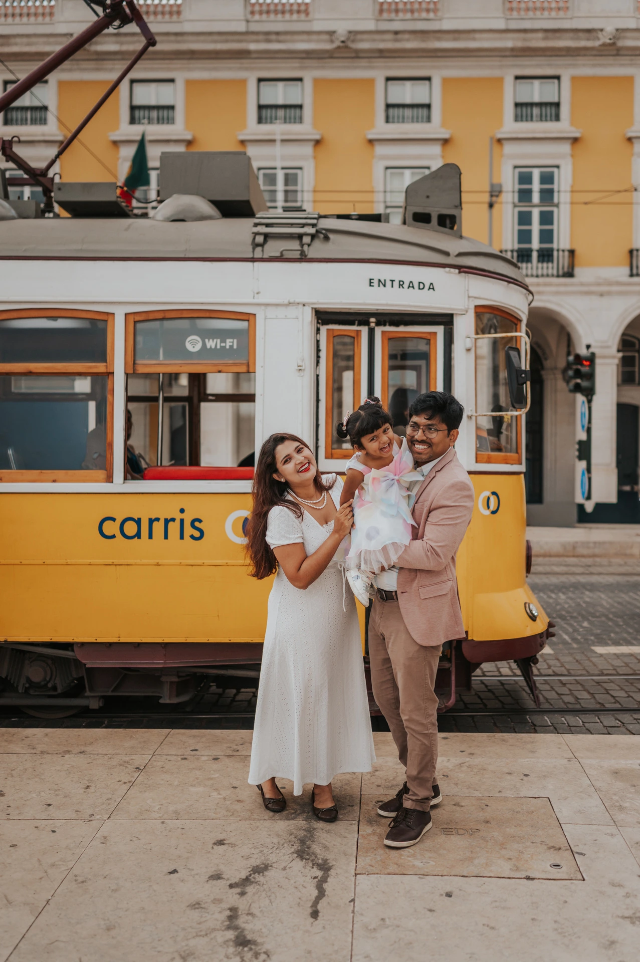 The parents holding their smiling daughter together beside a yellow Lisbon tram during their family birthday photoshoot in Lisbon.