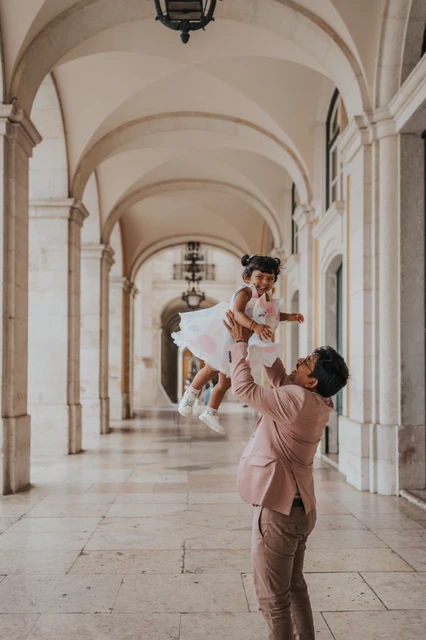 A playful birthday lift with her father | Lisbon family birthday photoshoot The father lifting his daughter into the air beneath the stone arcade during their family birthday photoshoot in Lisbon.