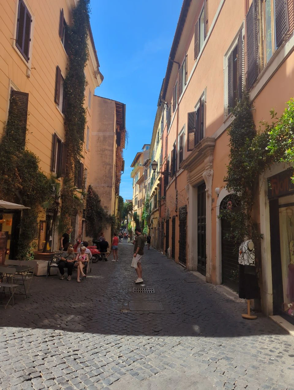 Trastevere Photoshoot - Lively Cobbled Street Visitors pause near café tables and leafy facades on a shaded cobbled street in Trastevere, Rome.