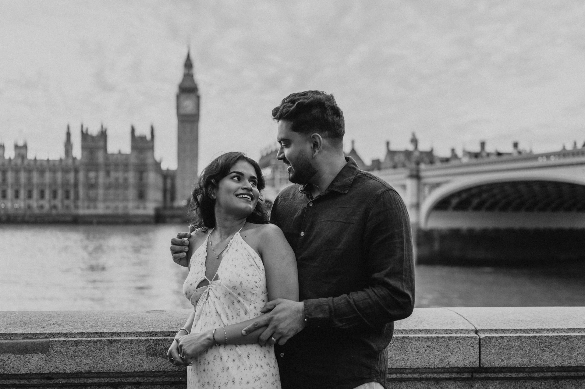 Black-and-white portrait of the couple looking at each other beside the Thames, with Big Ben behind them, from their London couple photoshoot.