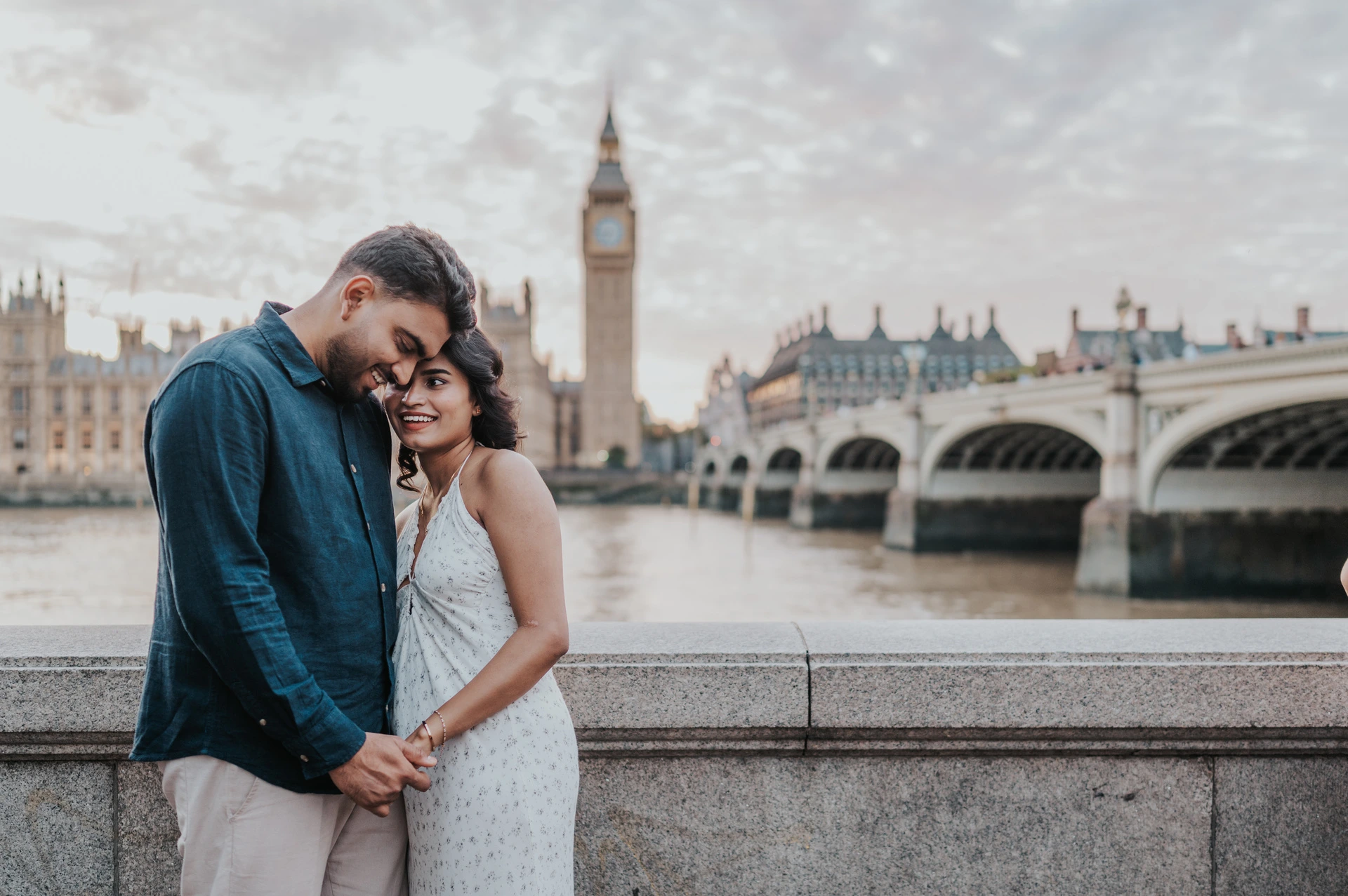Couple leaning their heads together beside the stone riverside wall, with Big Ben and the arches of Westminster Bridge behind, during their London couple photoshoot.