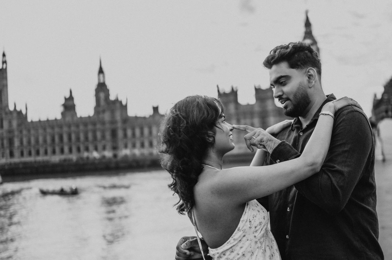 Black-and-white London couple photoshoot overlooking Parliament Black-and-white landscape portrait of the couple in an embrace beside Westminster Bridge’s parapet, with the Thames and Parliament behind, from their London couple photoshoot.
