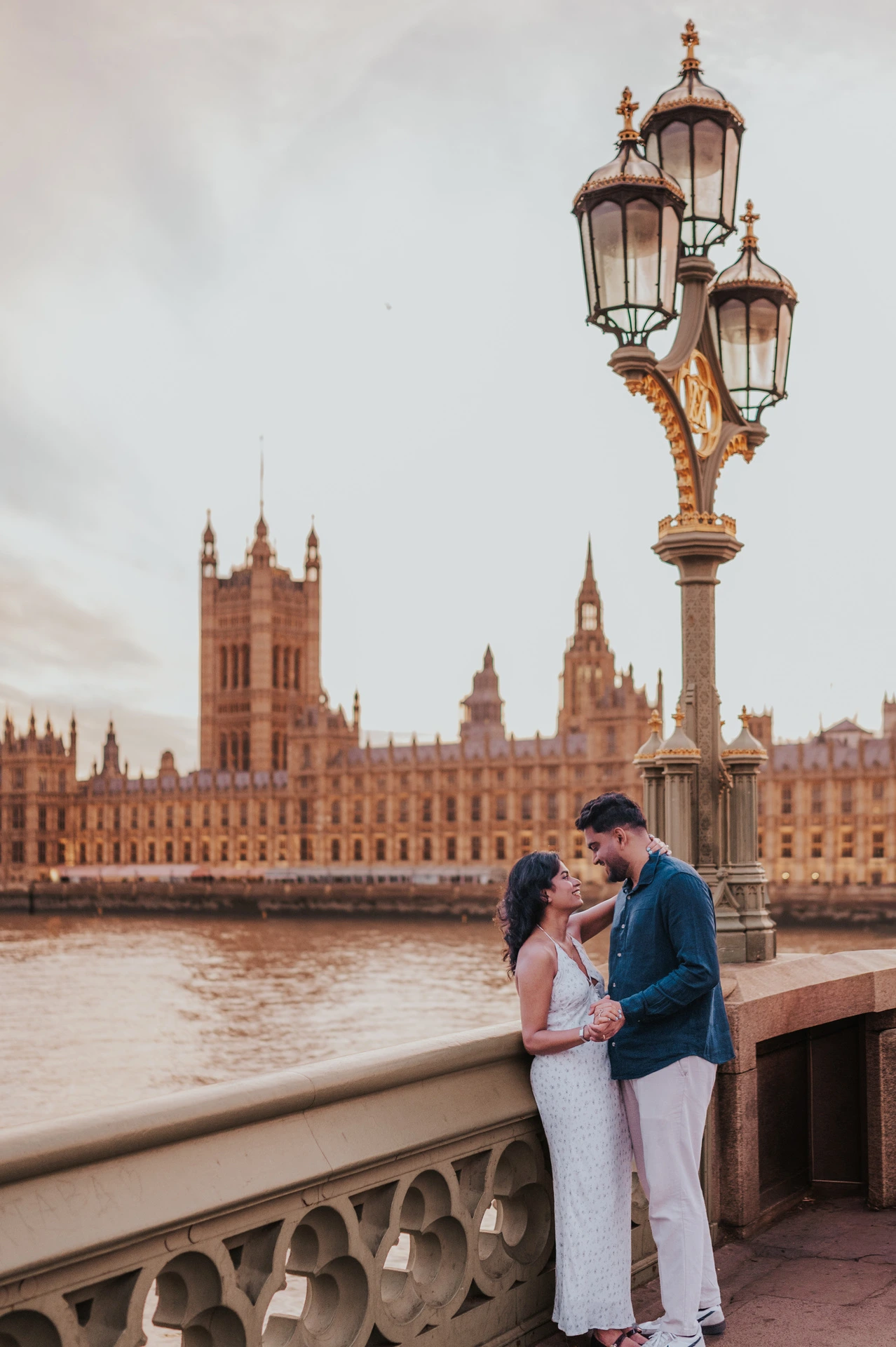 Full-length view of the couple holding hands beneath an ornate Westminster Bridge lamp, with the Houses of Parliament across the Thames, during their London couple photoshoot.
