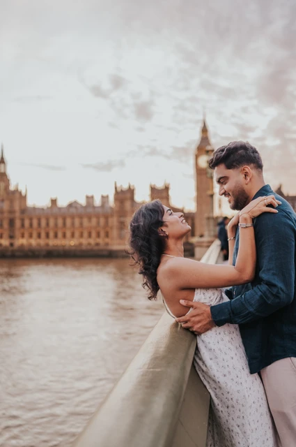 Couple facing each other in an embrace beside the Westminster Bridge parapet, with the Houses of Parliament behind, during their London couple photoshoot.