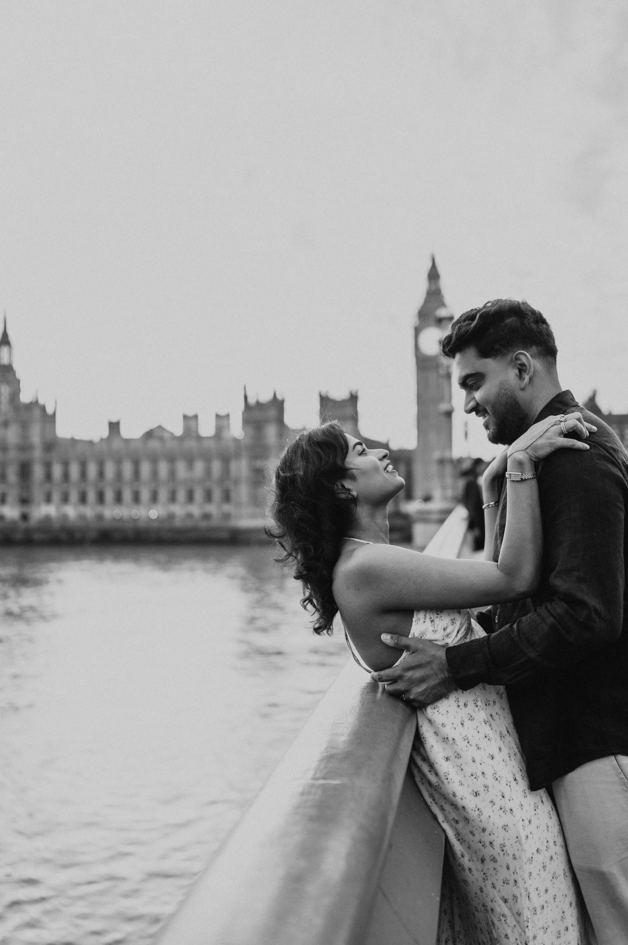 Black-and-white portrait of the woman with her arms around her partner’s shoulders at the Westminster Bridge parapet, from their London couple photoshoot.