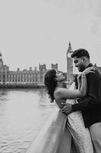 London couple photoshoot embracing on Westminster Bridge in black and white Black-and-white portrait of the woman with her arms around her partner’s shoulders at the Westminster Bridge parapet, from their London couple photoshoot.