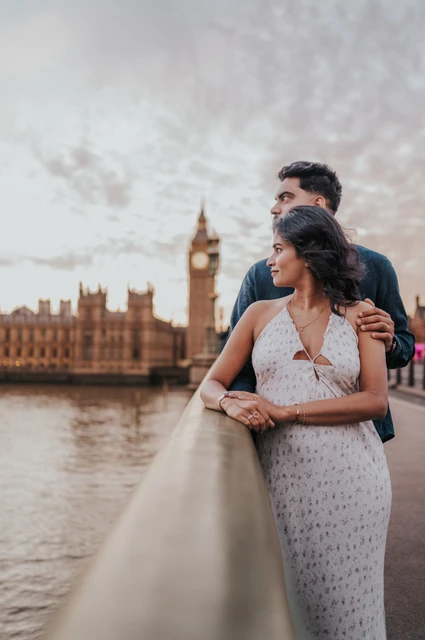 London couple photoshoot looking over the Thames from the bridge Couple looking across the Thames from the Westminster Bridge parapet, with Big Ben beyond the water, during their London couple photoshoot.