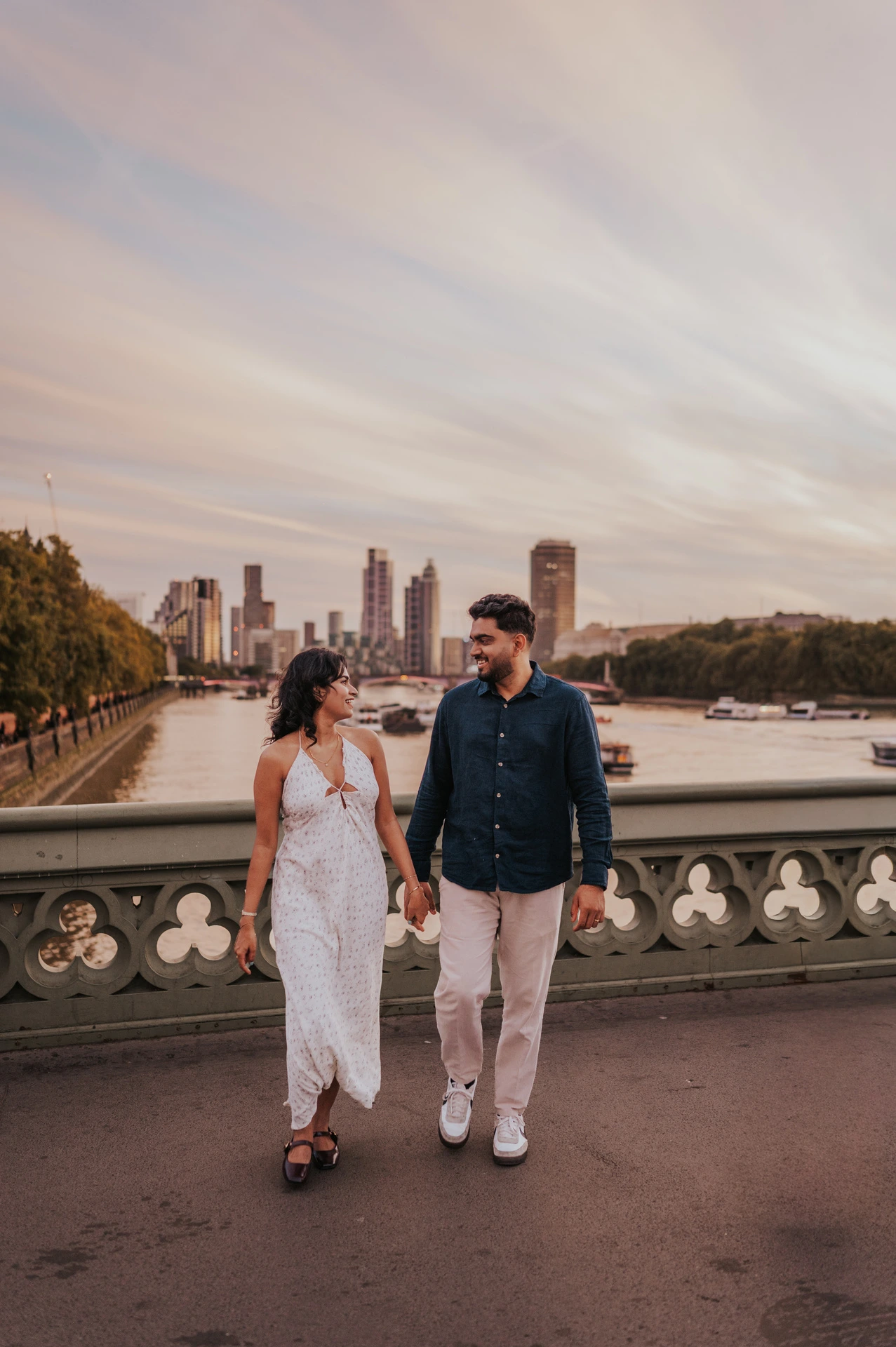 Couple walking hand in hand on Westminster Bridge beneath a broad sky, with the Thames beyond the patterned parapet, during their London couple photoshoot.