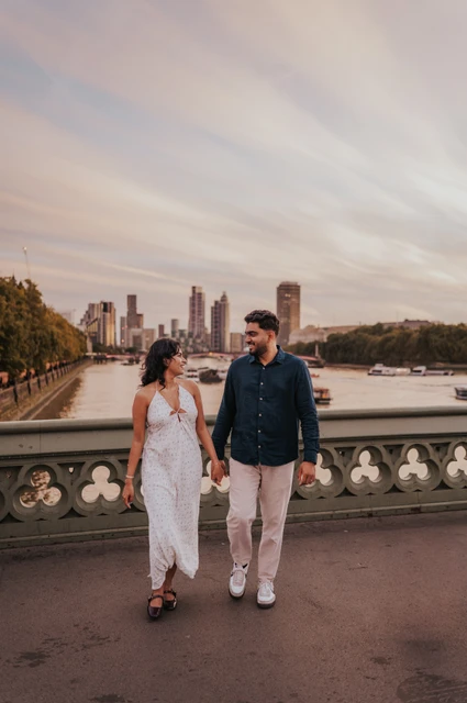 London couple photoshoot walking beneath the Westminster sky Couple walking hand in hand on Westminster Bridge beneath a broad sky, with the Thames beyond the patterned parapet, during their London couple photoshoot.