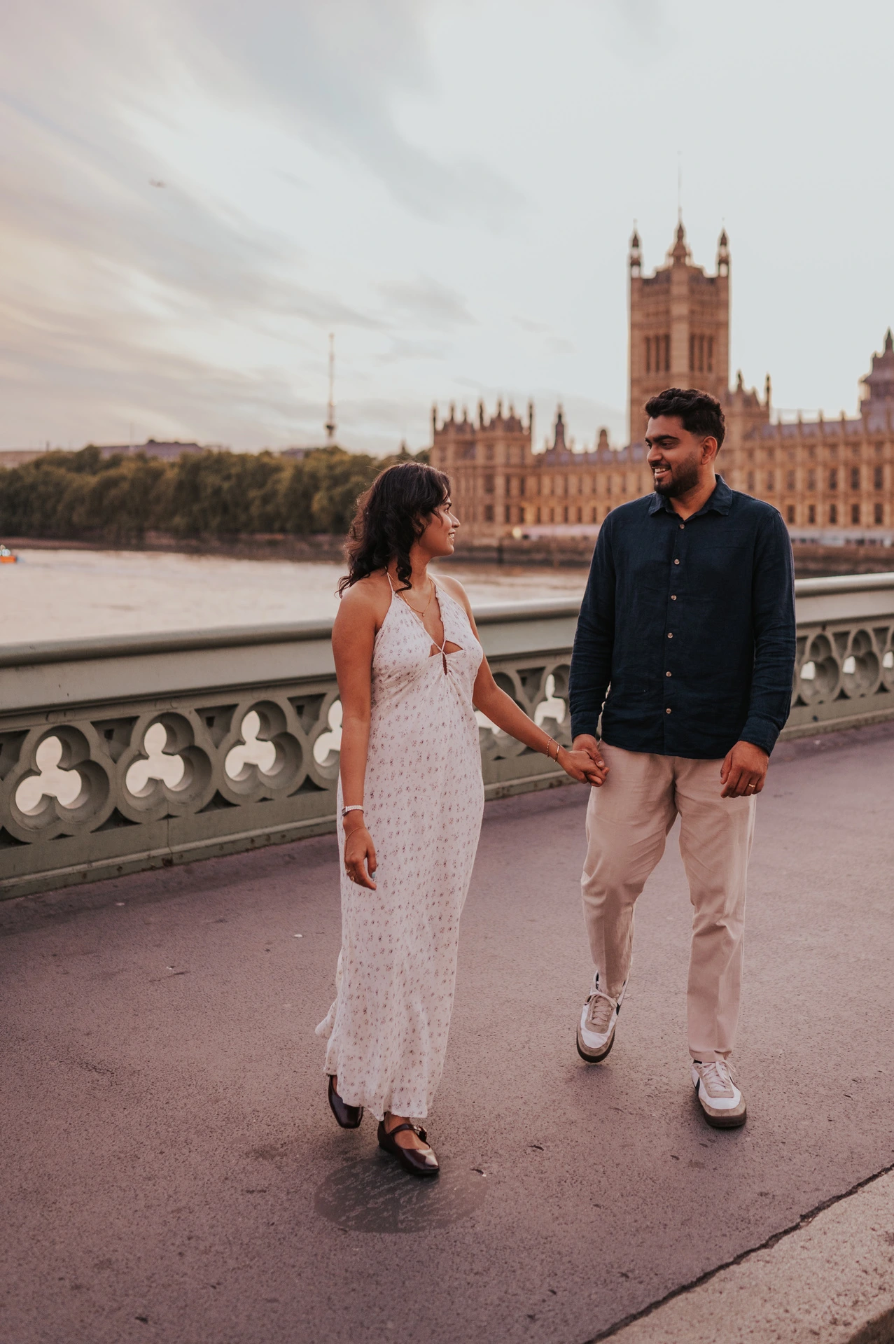 Couple walking hand in hand and looking at each other on Westminster Bridge, with Parliament beyond the parapet, during their London couple photoshoot.