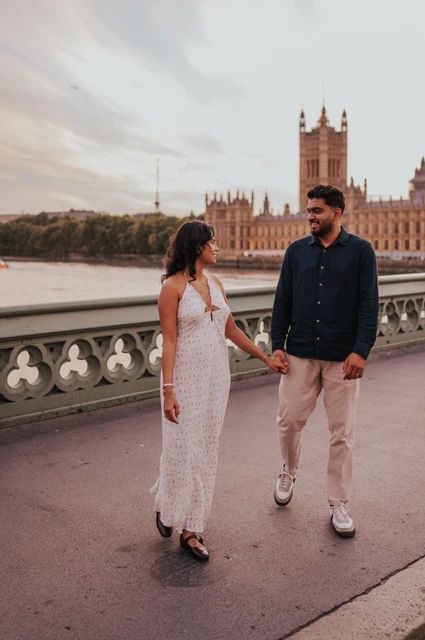 London couple photoshoot walking together on Westminster Bridge Couple walking hand in hand and looking at each other on Westminster Bridge, with Parliament beyond the parapet, during their London couple photoshoot.
