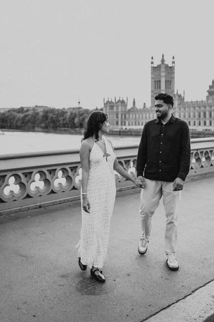 Black-and-white London couple photoshoot walking on the bridge Black-and-white photograph of the couple walking hand in hand on Westminster Bridge and looking at one another, from their London couple photoshoot.
