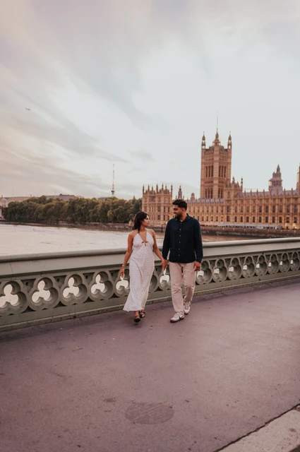 Wide view of the couple walking hand in hand on Westminster Bridge, with the patterned parapet and Houses of Parliament behind, during their London couple photoshoot.