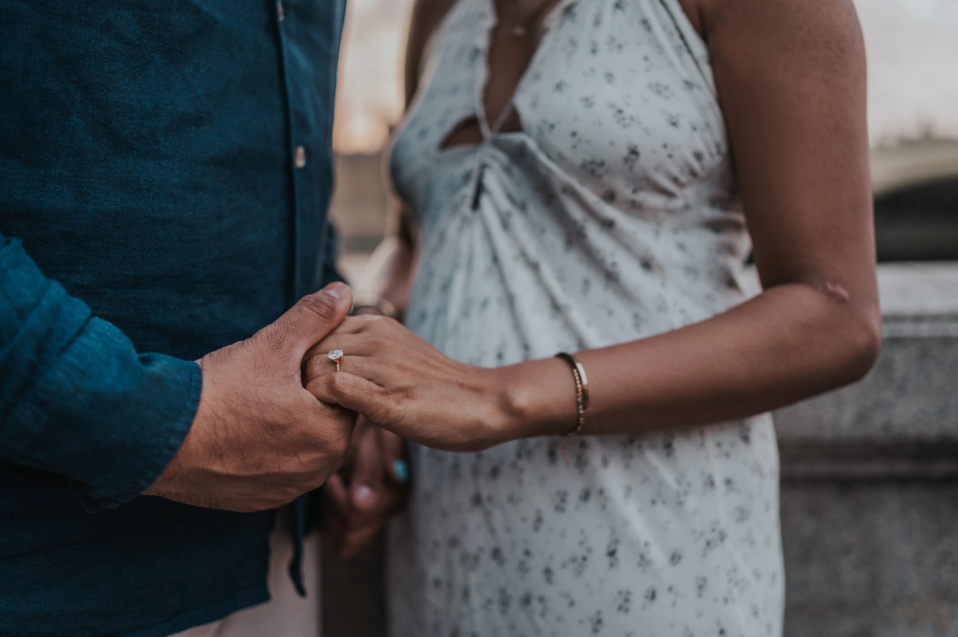 Close view of the couple’s clasped hands against her pale floral dress and his deep blue shirt, from their London couple photoshoot.