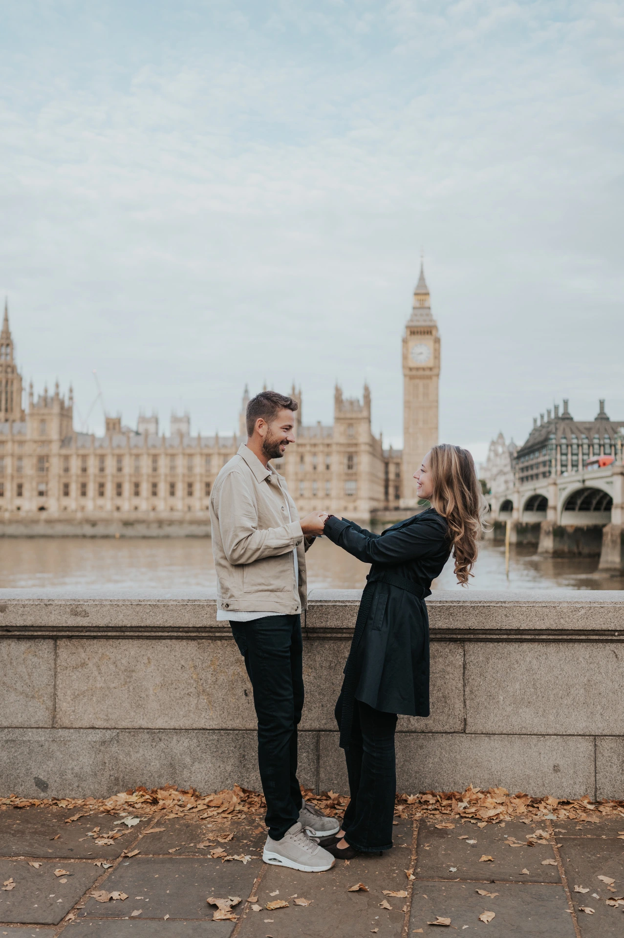 Full-length portrait of a couple holding hands beside the Thames, with Big Ben and Westminster Bridge behind them, during their London photoshoot.