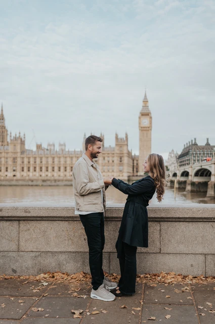 London couple photoshoot holding hands with Big Ben behind Full-length portrait of a couple holding hands beside the Thames, with Big Ben and Westminster Bridge behind them, during their London photoshoot.