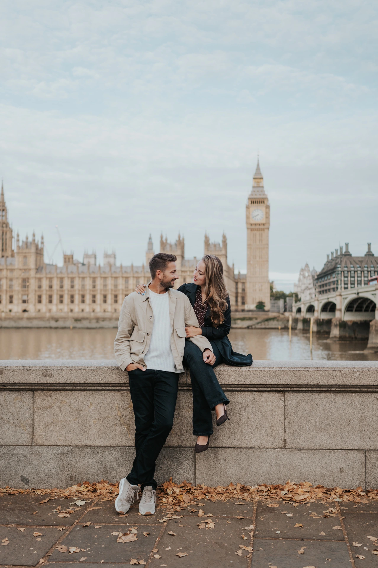 Couple looking at one another, with the woman seated on the riverside wall and Big Ben across the Thames, during their London couple photoshoot.