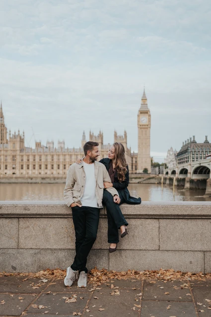 London couple photoshoot on the riverside wall by Big Ben Couple looking at one another, with the woman seated on the riverside wall and Big Ben across the Thames, during their London couple photoshoot.