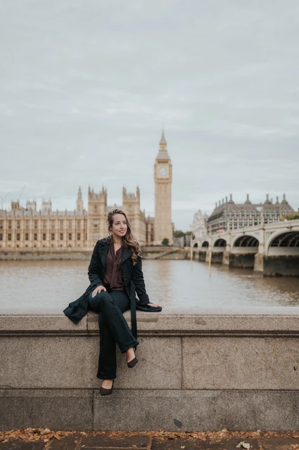 Woman facing the camera while seated on the stone riverside wall, with Big Ben behind her, during a London couple photoshoot.