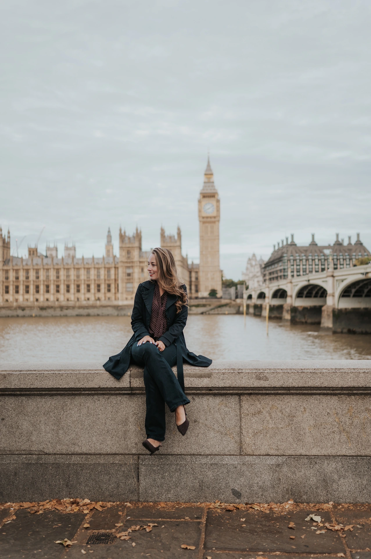 Woman in a dark coat seated on the riverside wall, looking to the side with Big Ben across the Thames during a London couple photoshoot.