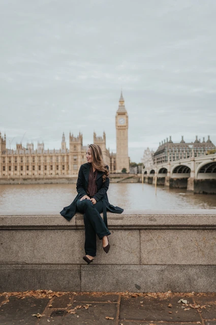 London photoshoot with a solo portrait beside the Thames Woman in a dark coat seated on the riverside wall, looking to the side with Big Ben across the Thames during a London couple photoshoot.