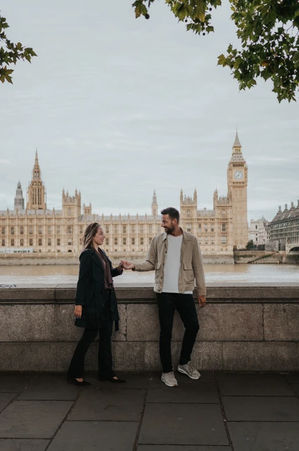 London couple photoshoot beneath trees with Big Ben behind Couple holding hands beneath the riverside trees, with Big Ben and Parliament across the Thames, during their London couple photoshoot.