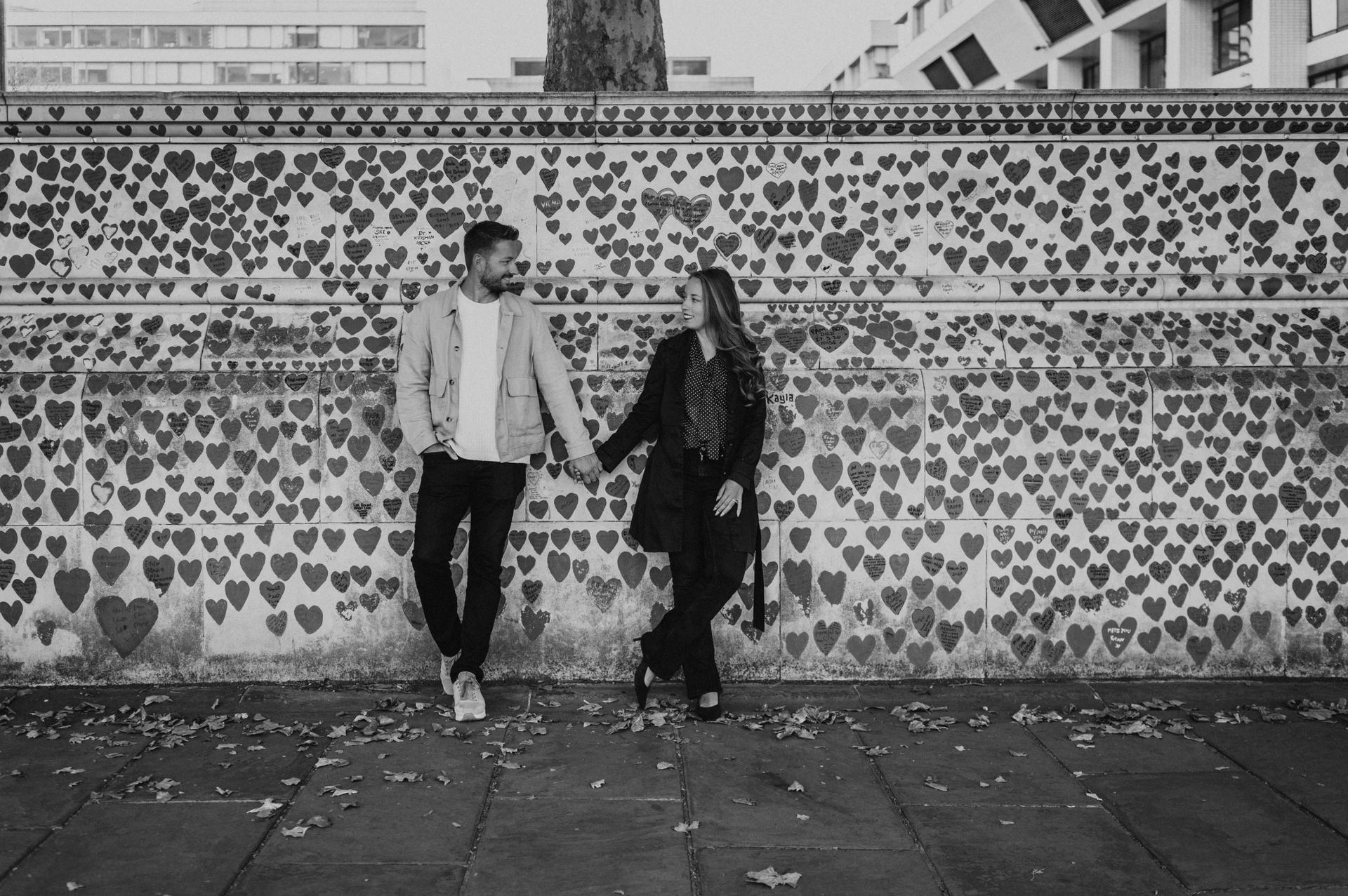 Black-and-white wide portrait of a couple holding hands beside the National Covid Memorial Wall during their London couple photoshoot.