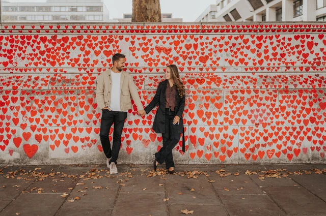 Wide portrait of a couple holding hands beside the National Covid Memorial Wall, with fallen leaves on the pavement, during their London photoshoot.