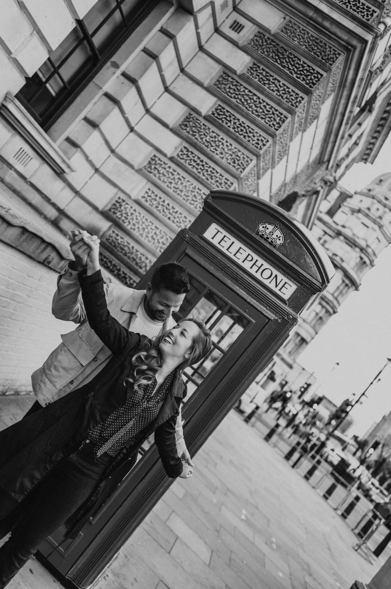 Black-and-white portrait of a smiling couple with their joined hands raised beside a telephone box during their London photoshoot.