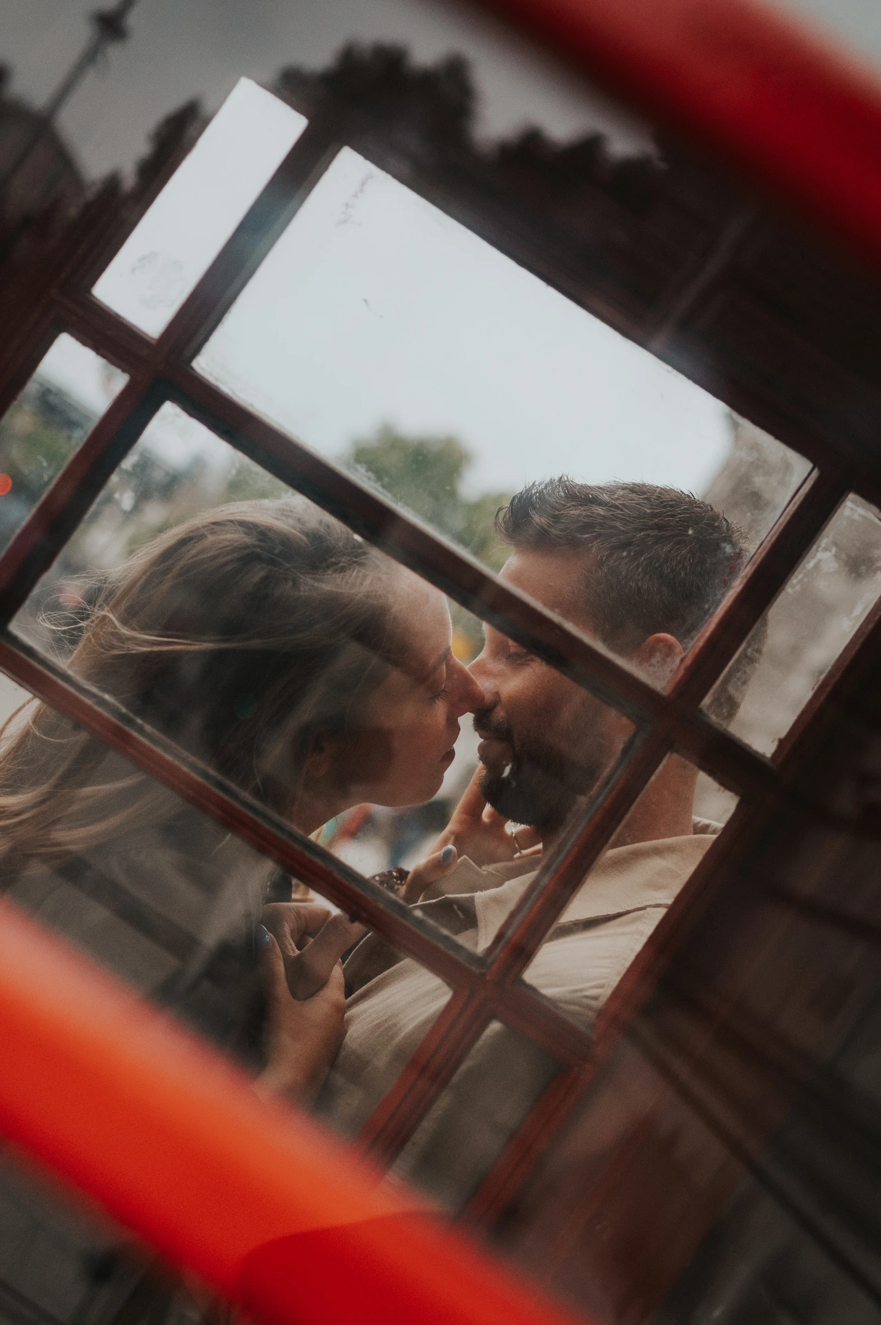 Close view of a couple kissing through the red-framed glass panes of a telephone box during their London couple photoshoot.