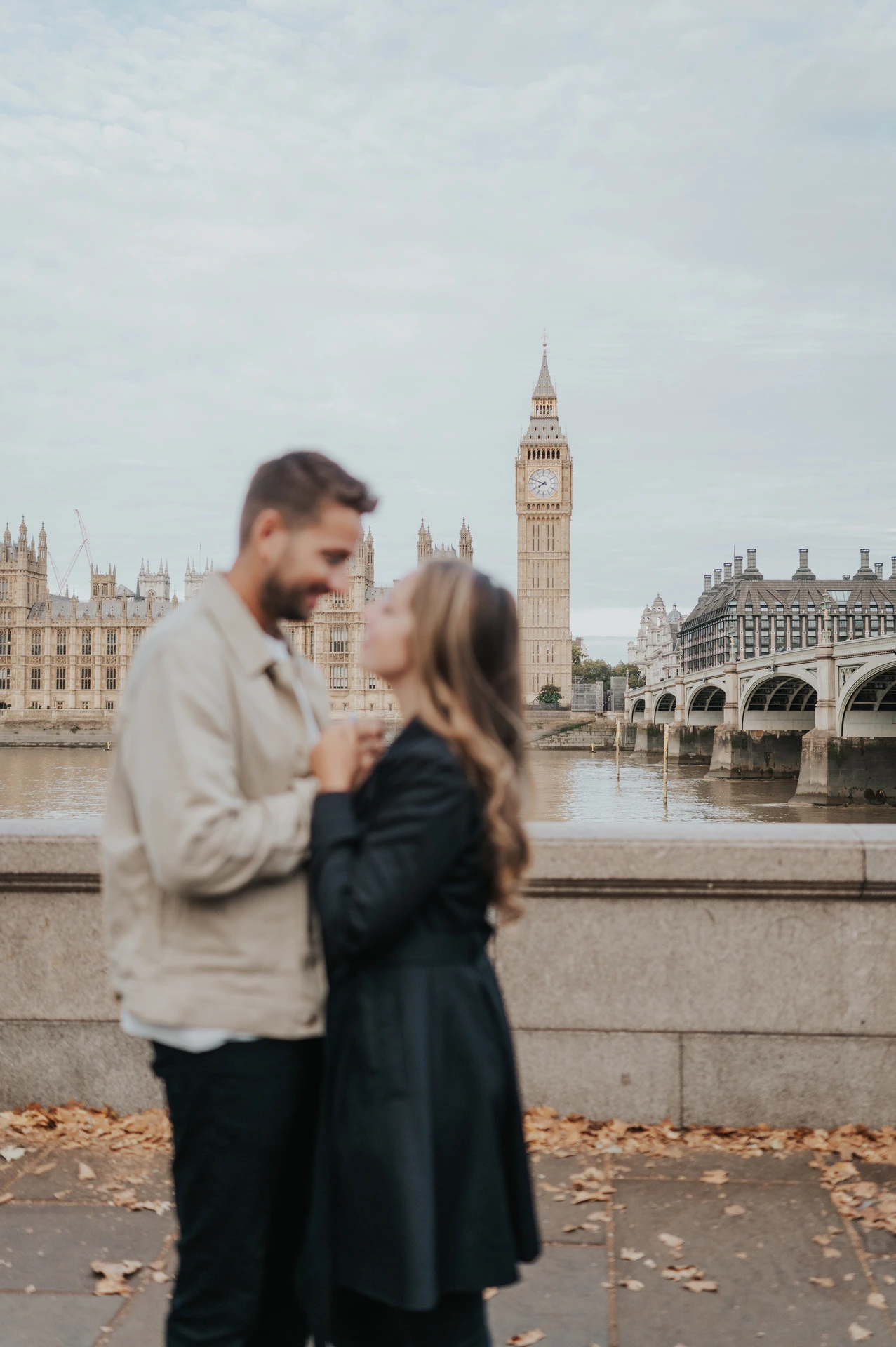 Couple looking at one another in a medium portrait beside the Thames, with her hands resting on his light jacket, during their London photoshoot.