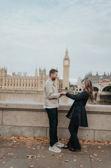 London couple photoshoot smiling by the riverside wall A woman in a dark coat smiling towards her partner beside the riverside wall, with Big Ben across the Thames during their London couple photoshoot.